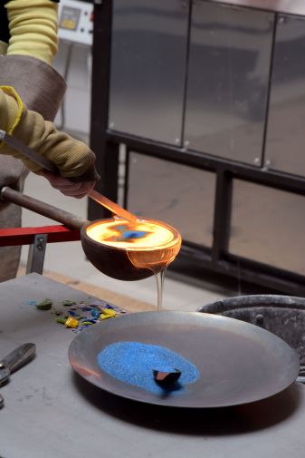 Arbeit am Studioofen beim Internationalen Glassymposium 2025 an der Glasfachschule NRW, geschmolzenes Glas in einer Kelle wird mit blauen Farbpigmenten eingefärbt, blaue Glasstücke auf dem Tisch