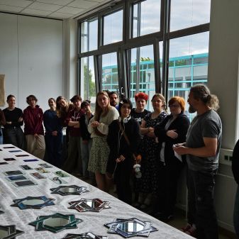 Eine Gruppe von Schülerinnen und Schülern steht in einem Raum mit großen Fenstern und betrachtet einen Tisch, der mit verschiedenen geometrisch geformten Spiegeln bedeckt ist.