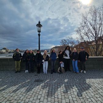 Gruppe von neun Personen steht auf der Karlsbrücke in Prag vor einer Laterne mit Fluss und Gebäuden im Hintergrund bei bewölktem Himmel.